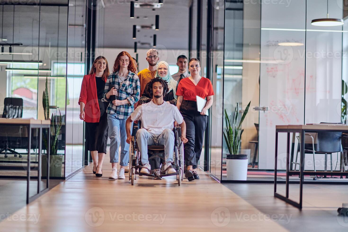 A diverse group of young business people congregates within a modern startup's glass-enclosed office, featuring inclusivity with a person in a wheelchair, an African American young man , and a hijab photo