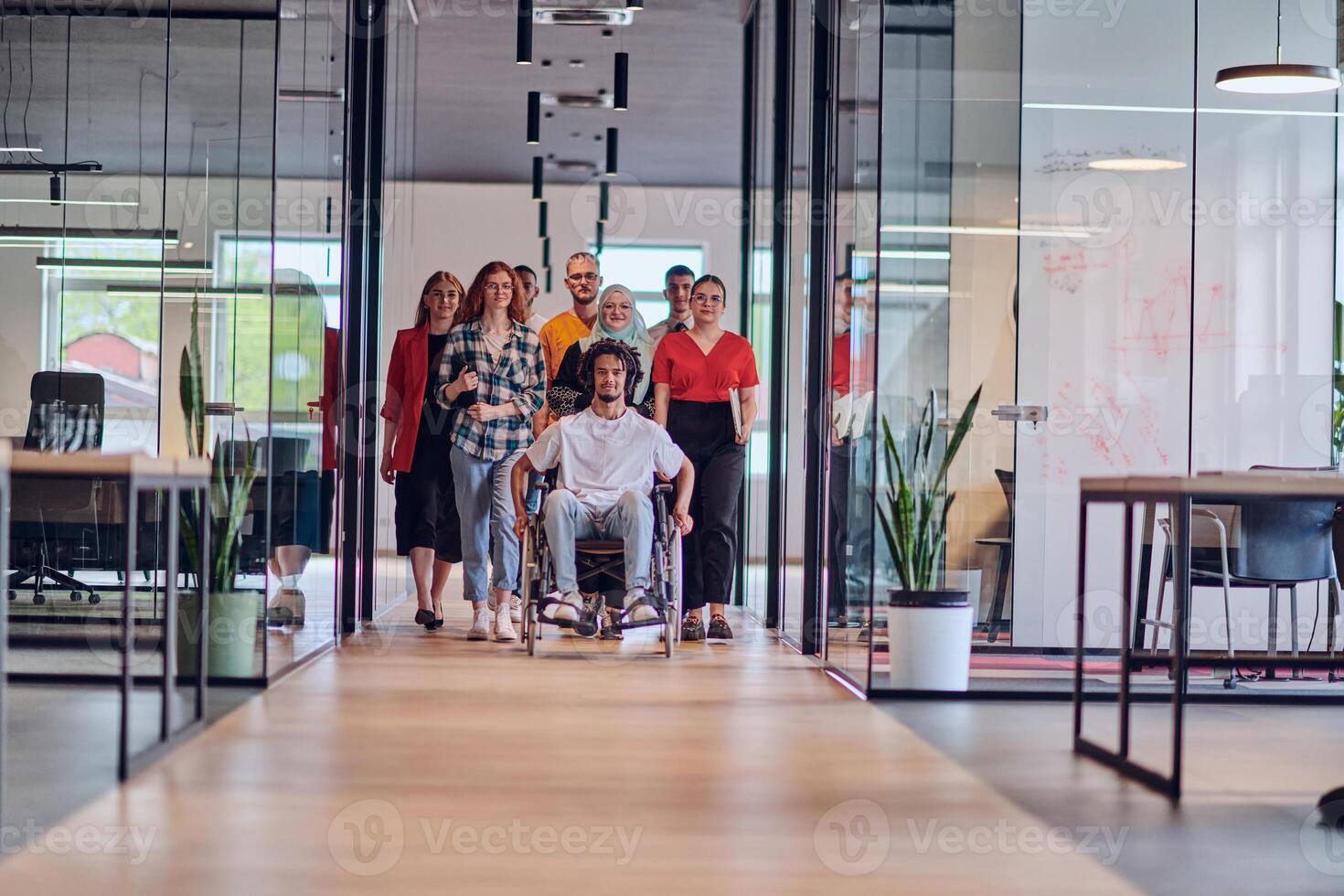 A diverse group of young business people congregates within a modern startup's glass-enclosed office, featuring inclusivity with a person in a wheelchair, an African American young man , and a hijab photo