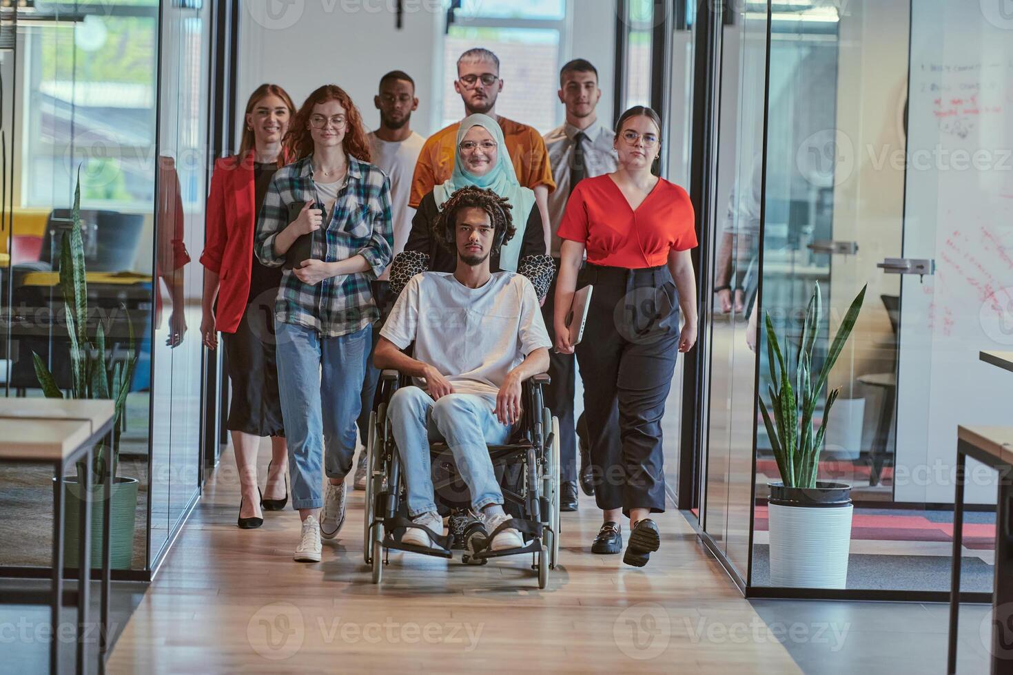 A diverse group of young business people walking a corridor in the glass-enclosed office of a modern startup, including a person in a wheelchair and a woman wearing a hijab, showing a dynamic mix of photo