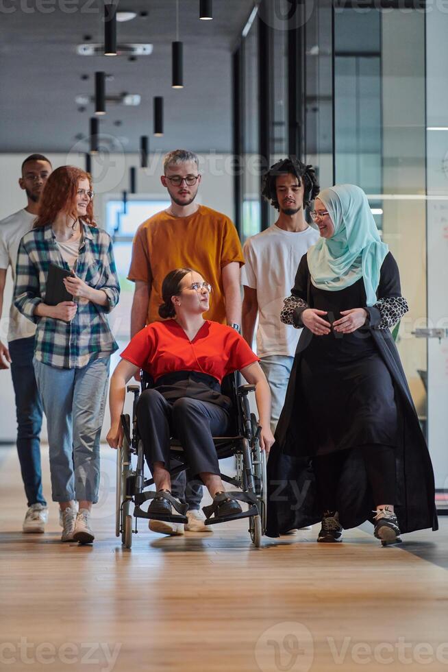 A diverse group of young business people walking a corridor in the glass-enclosed office of a modern startup, including a person in a wheelchair and a woman wearing a hijab, showing a dynamic mix of photo