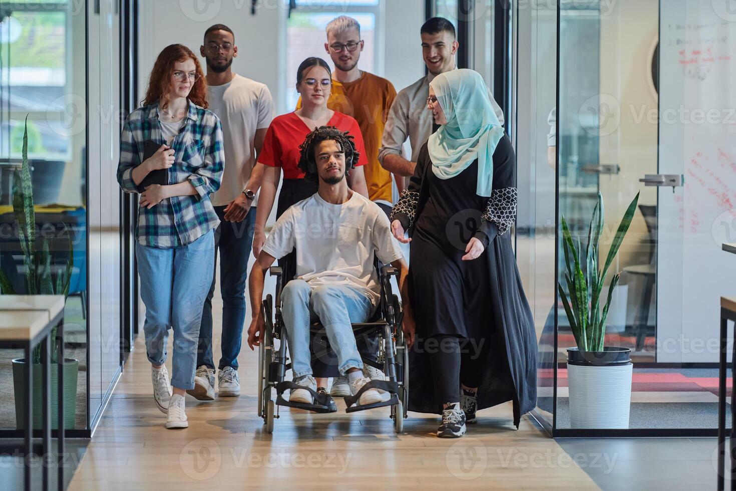 A diverse group of young business people walking a corridor in the glass-enclosed office of a modern startup, including a person in a wheelchair and a woman wearing a hijab, showing a dynamic mix of photo