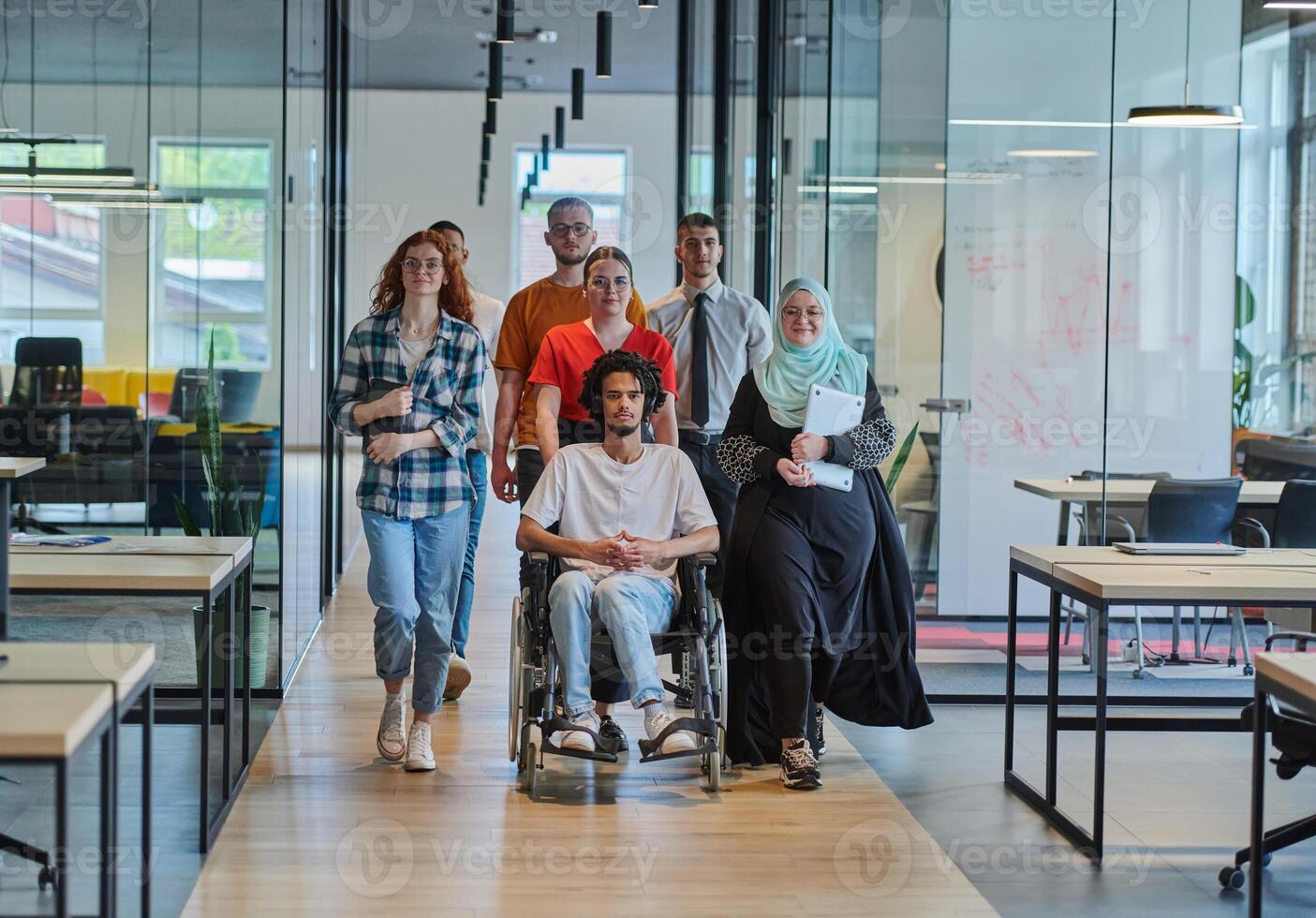 A diverse group of young business people walking a corridor in the glass-enclosed office of a modern startup, including a person in a wheelchair and a woman wearing a hijab, showing a dynamic mix of photo
