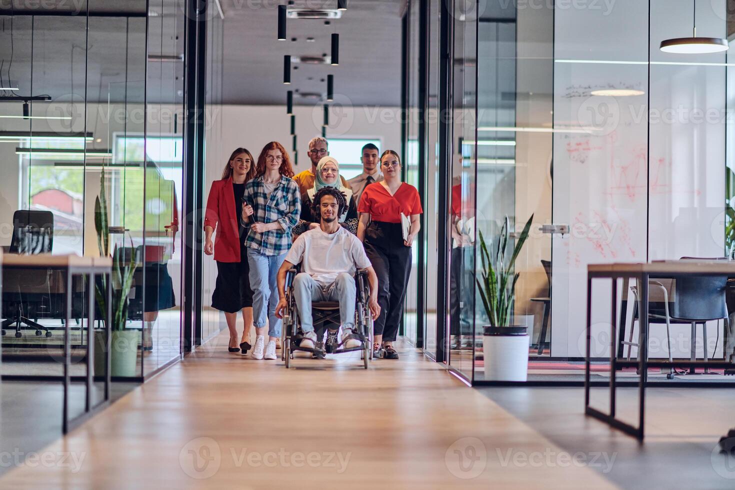 A diverse group of young business people congregates within a modern startup's glass-enclosed office, featuring inclusivity with a person in a wheelchair, an African American young man , and a hijab photo