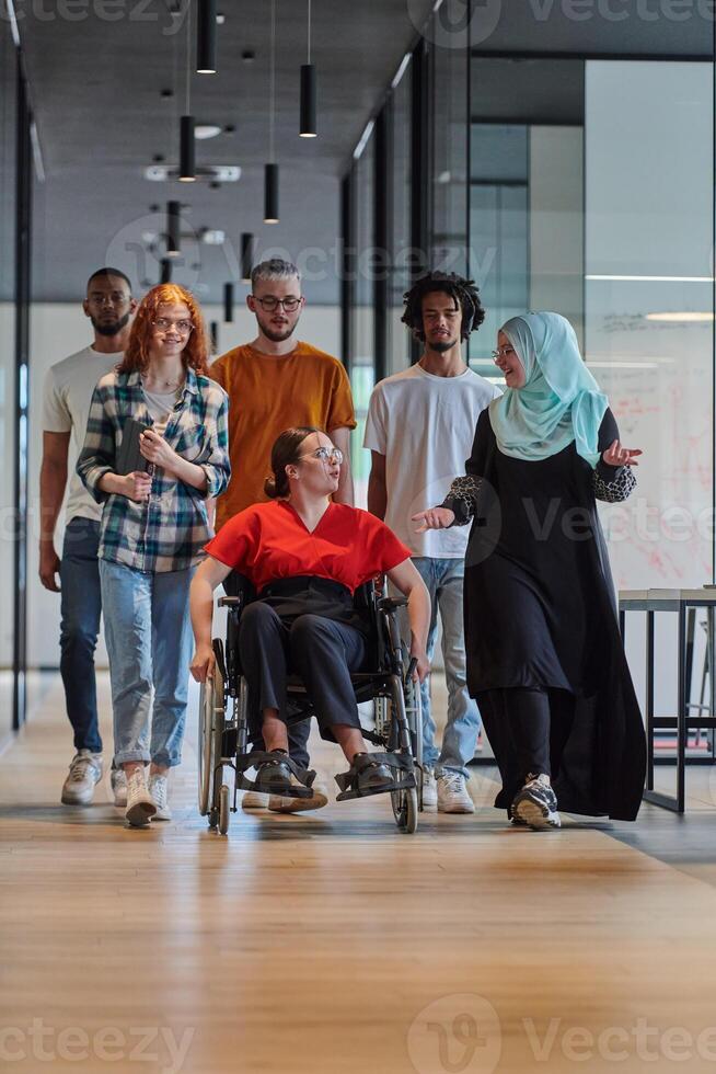 A diverse group of young business people walking a corridor in the glass-enclosed office of a modern startup, including a person in a wheelchair and a woman wearing a hijab, showing a dynamic mix of photo
