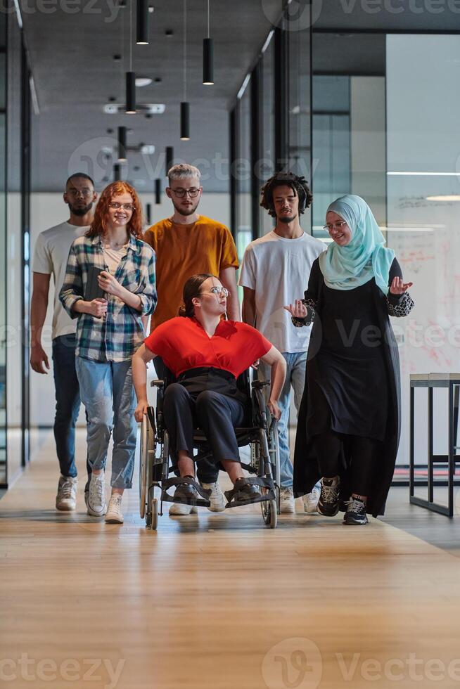 A diverse group of young business people walking a corridor in the glass-enclosed office of a modern startup, including a person in a wheelchair and a woman wearing a hijab, showing a dynamic mix of photo