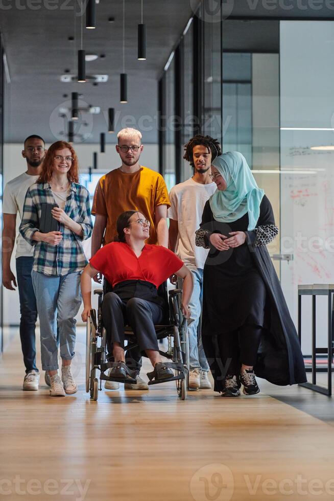 A diverse group of young business people walking a corridor in the glass-enclosed office of a modern startup, including a person in a wheelchair and a woman wearing a hijab, showing a dynamic mix of photo