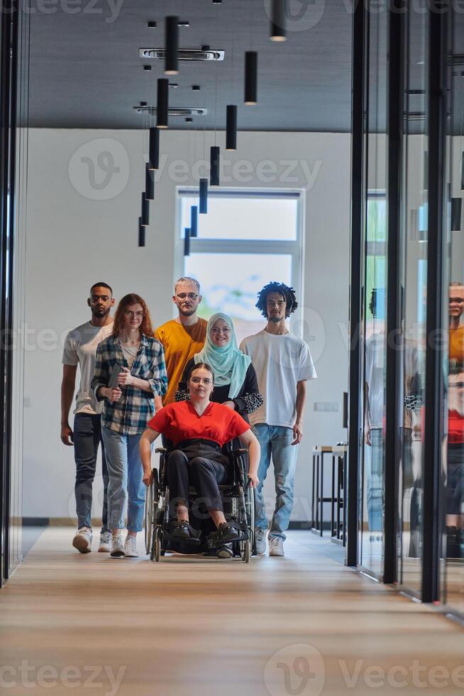 A diverse group of young business people walking a corridor in the glass-enclosed office of a modern startup, including a person in a wheelchair and a woman wearing a hijab, showing a dynamic mix of photo