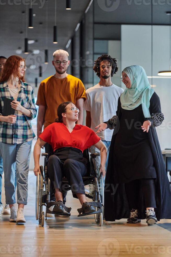 A diverse group of young business people walking a corridor in the glass-enclosed office of a modern startup, including a person in a wheelchair and a woman wearing a hijab, showing a dynamic mix of photo