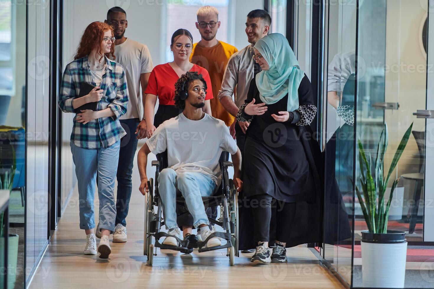 A diverse group of young business people walking a corridor in the glass-enclosed office of a modern startup, including a person in a wheelchair and a woman wearing a hijab, showing a dynamic mix of photo