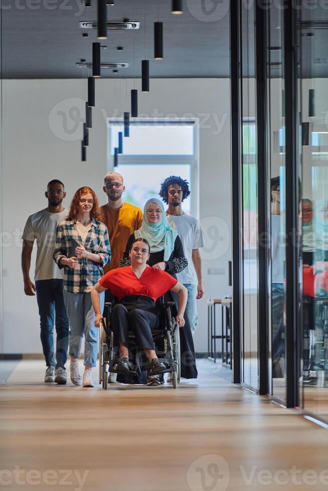 A diverse group of young business people walking a corridor in the glass-enclosed office of a modern startup, including a person in a wheelchair and a woman wearing a hijab, showing a dynamic mix of photo