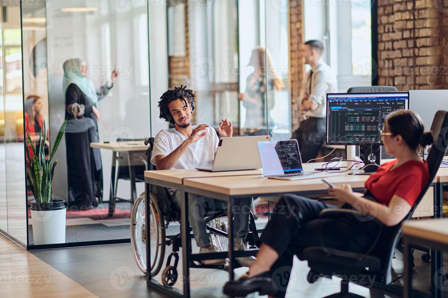 A young business group, including an African American businessman in a wheelchair, collaborates within a modern glass office, actively engaged around a computer and laptop, collectively solving photo