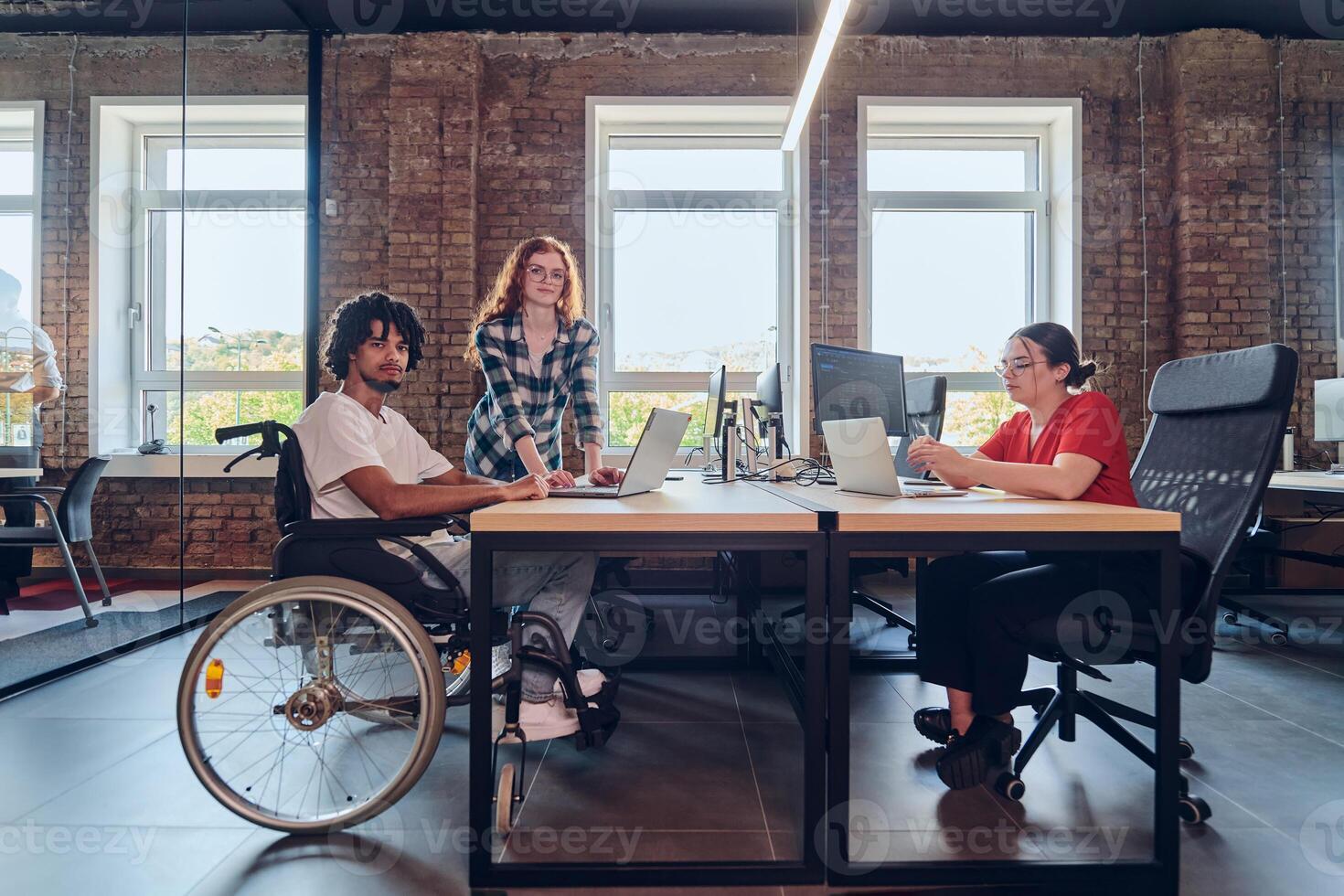 A young business group, including an African American businessman in a wheelchair, collaborates within a modern glass office, actively engaged around a computer and laptop, collectively solving photo