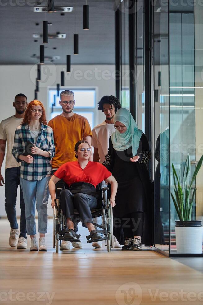 A diverse group of young business people walking a corridor in the glass-enclosed office of a modern startup, including a person in a wheelchair and a woman wearing a hijab, showing a dynamic mix of photo