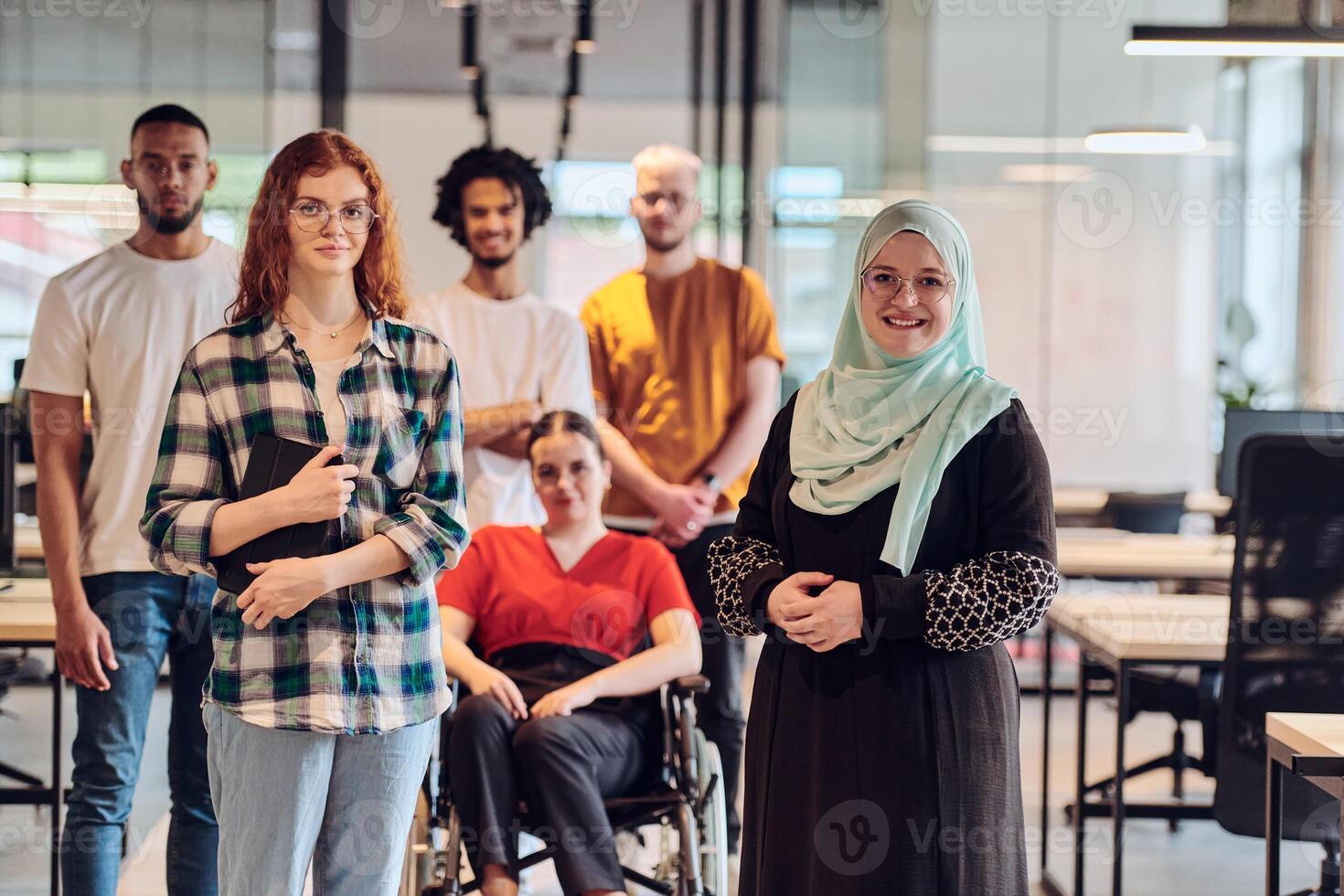A diverse group of young business people congregates within a modern startup's glass-enclosed office, featuring inclusivity with a person in a wheelchair, an African American young man , and a hijab photo