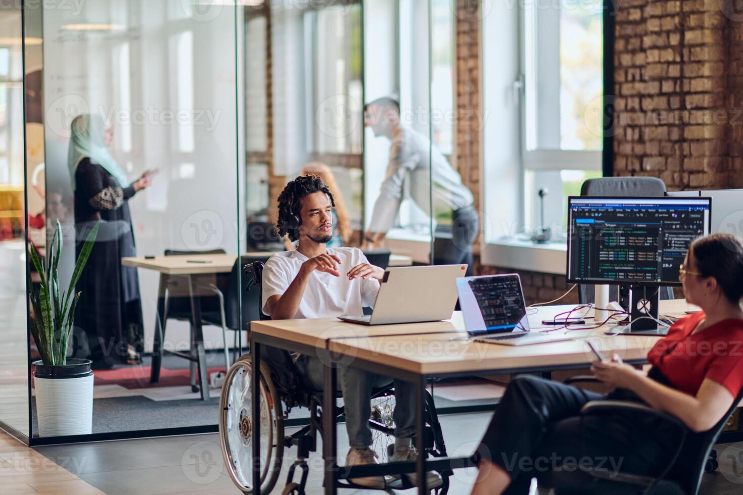 A young business group, including an African American businessman in a wheelchair, collaborates within a modern glass office, actively engaged around a computer and laptop, collectively solving photo