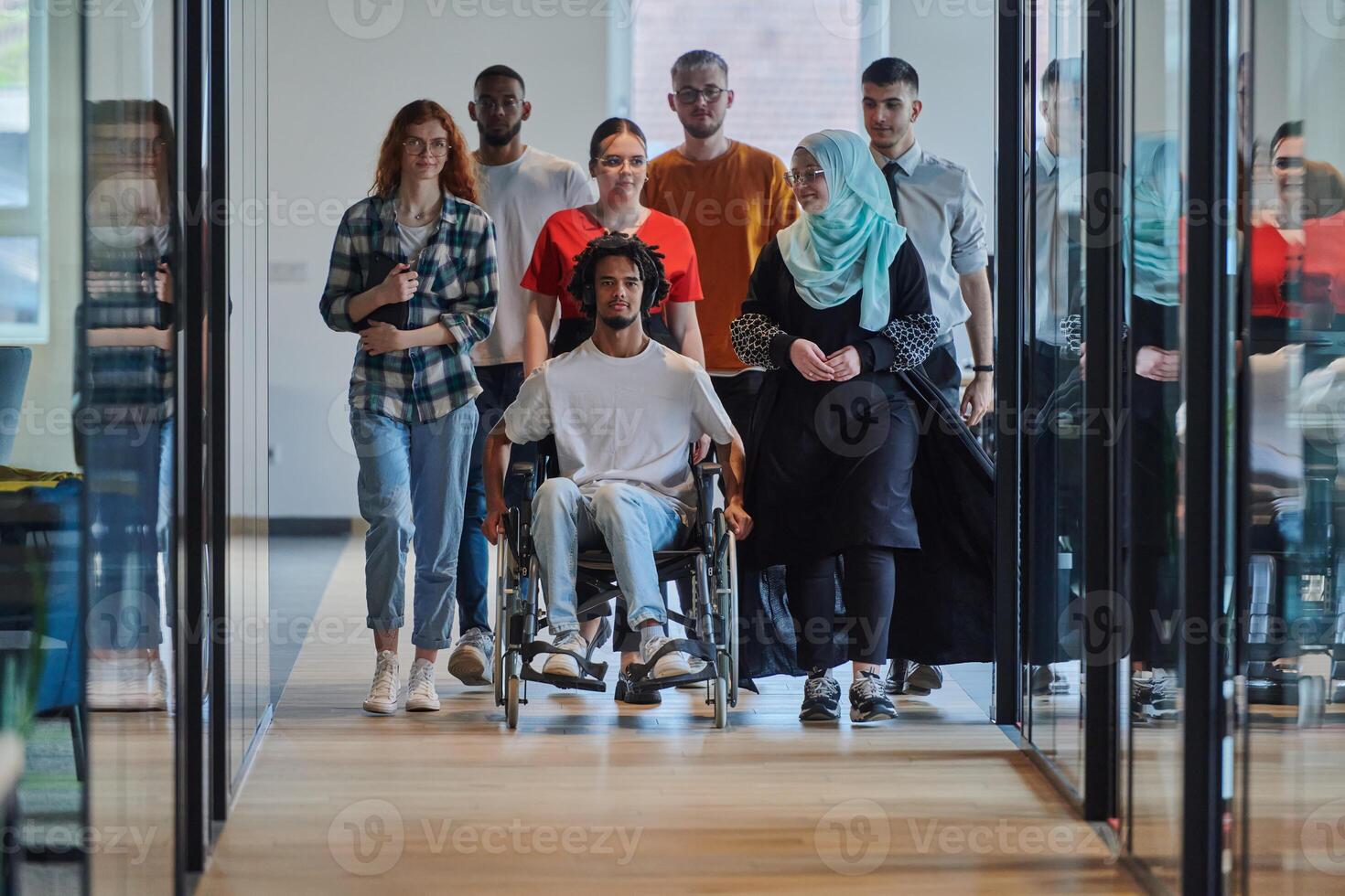 A diverse group of young business people walking a corridor in the glass-enclosed office of a modern startup, including a person in a wheelchair and a woman wearing a hijab, showing a dynamic mix of photo