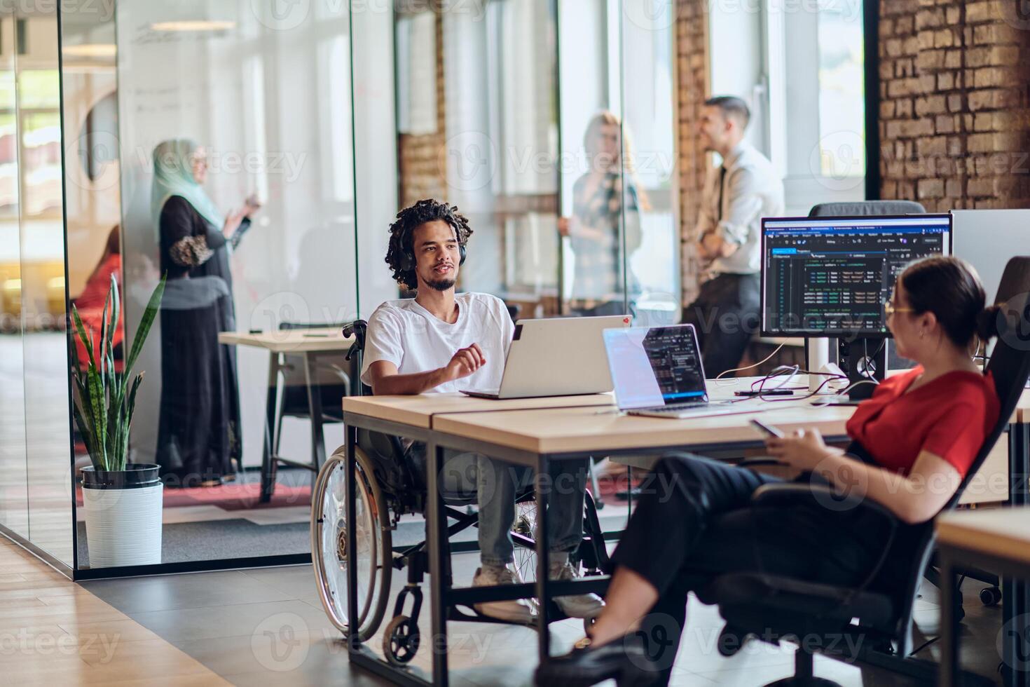 A young business group, including an African American businessman in a wheelchair, collaborates within a modern glass office, actively engaged around a computer and laptop, collectively solving photo