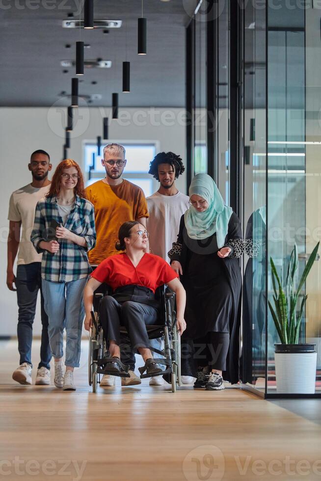 A diverse group of young business people walking a corridor in the glass-enclosed office of a modern startup, including a person in a wheelchair and a woman wearing a hijab, showing a dynamic mix of photo