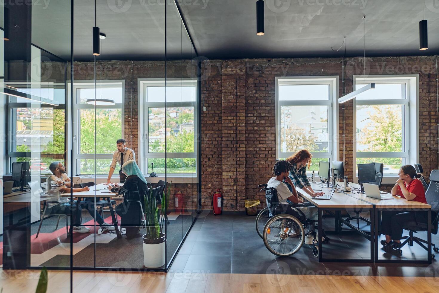 A young business group, including an African American businessman in a wheelchair, collaborates within a modern glass office, actively engaged around a computer and laptop, collectively solving photo