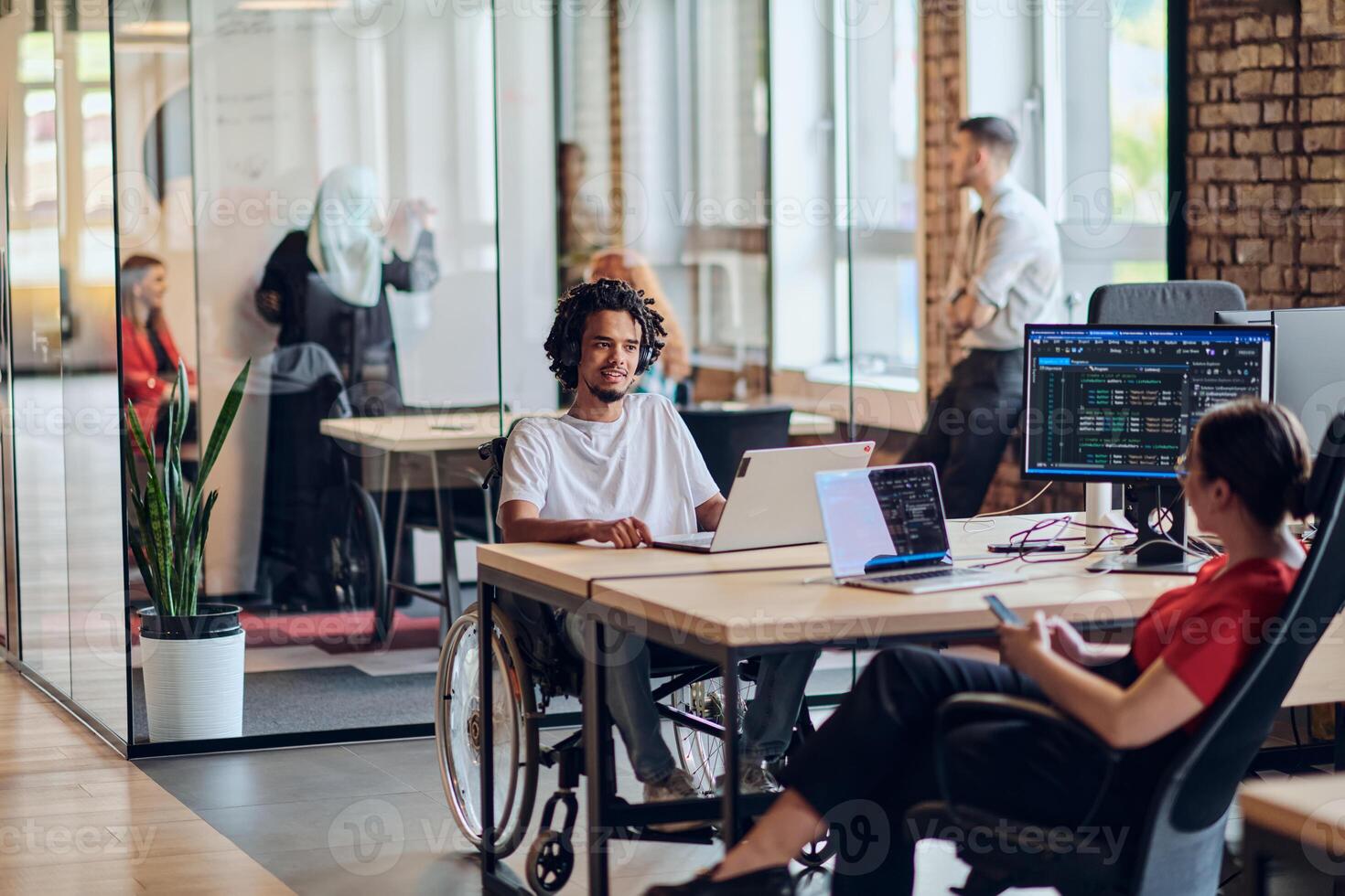 A young business group, including an African American businessman in a wheelchair, collaborates within a modern glass office, actively engaged around a computer and laptop, collectively solving photo