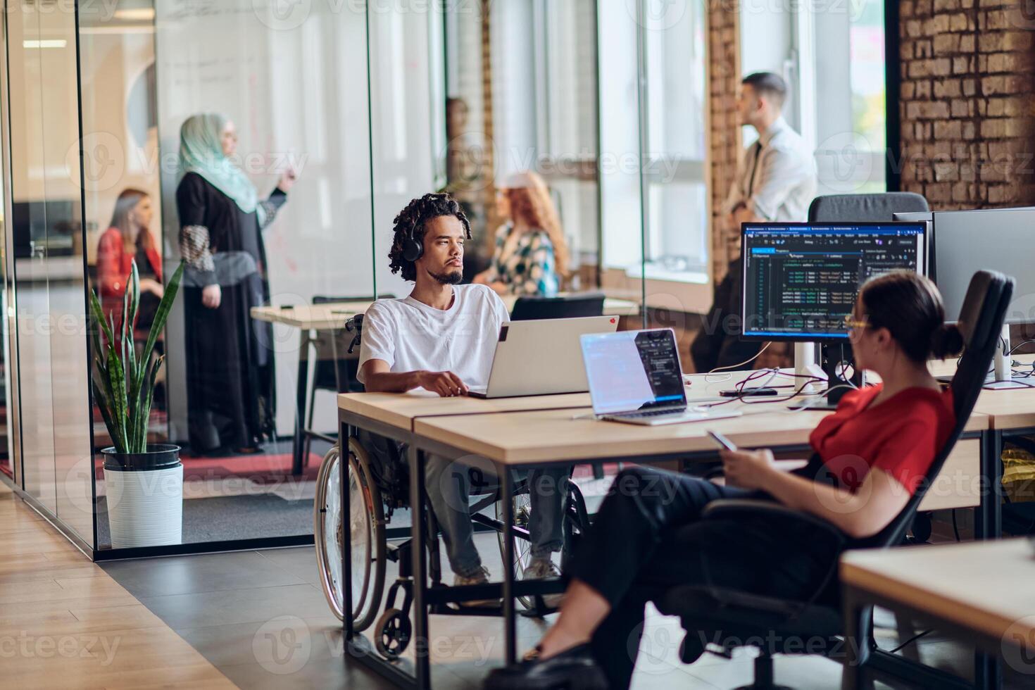 A young business group, including an African American businessman in a wheelchair, collaborates within a modern glass office, actively engaged around a computer and laptop, collectively solving photo