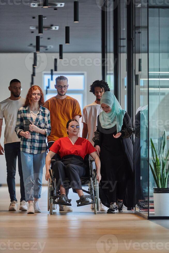A diverse group of young business people walking a corridor in the glass-enclosed office of a modern startup, including a person in a wheelchair and a woman wearing a hijab, showing a dynamic mix of photo