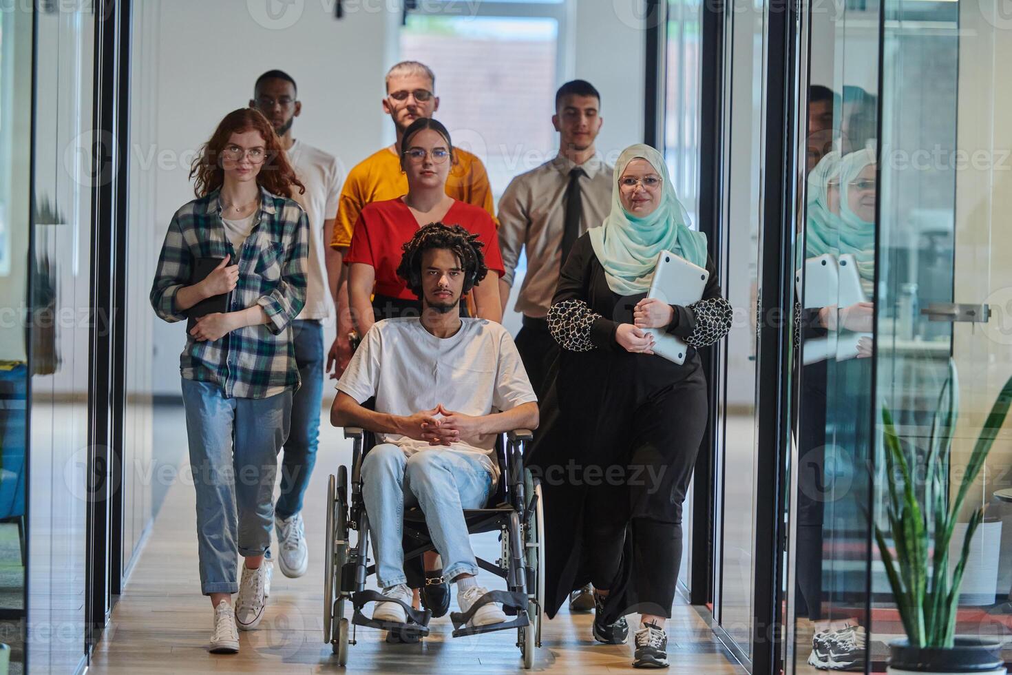 A diverse group of young business people walking a corridor in the glass-enclosed office of a modern startup, including a person in a wheelchair and a woman wearing a hijab, showing a dynamic mix of photo