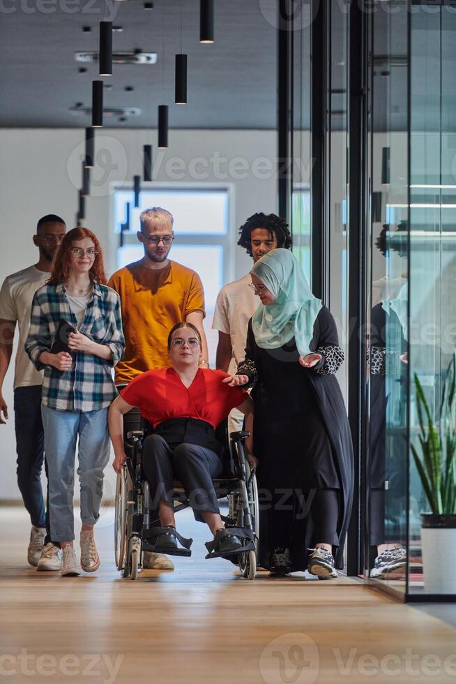 A diverse group of young business people walking a corridor in the glass-enclosed office of a modern startup, including a person in a wheelchair and a woman wearing a hijab, showing a dynamic mix of photo