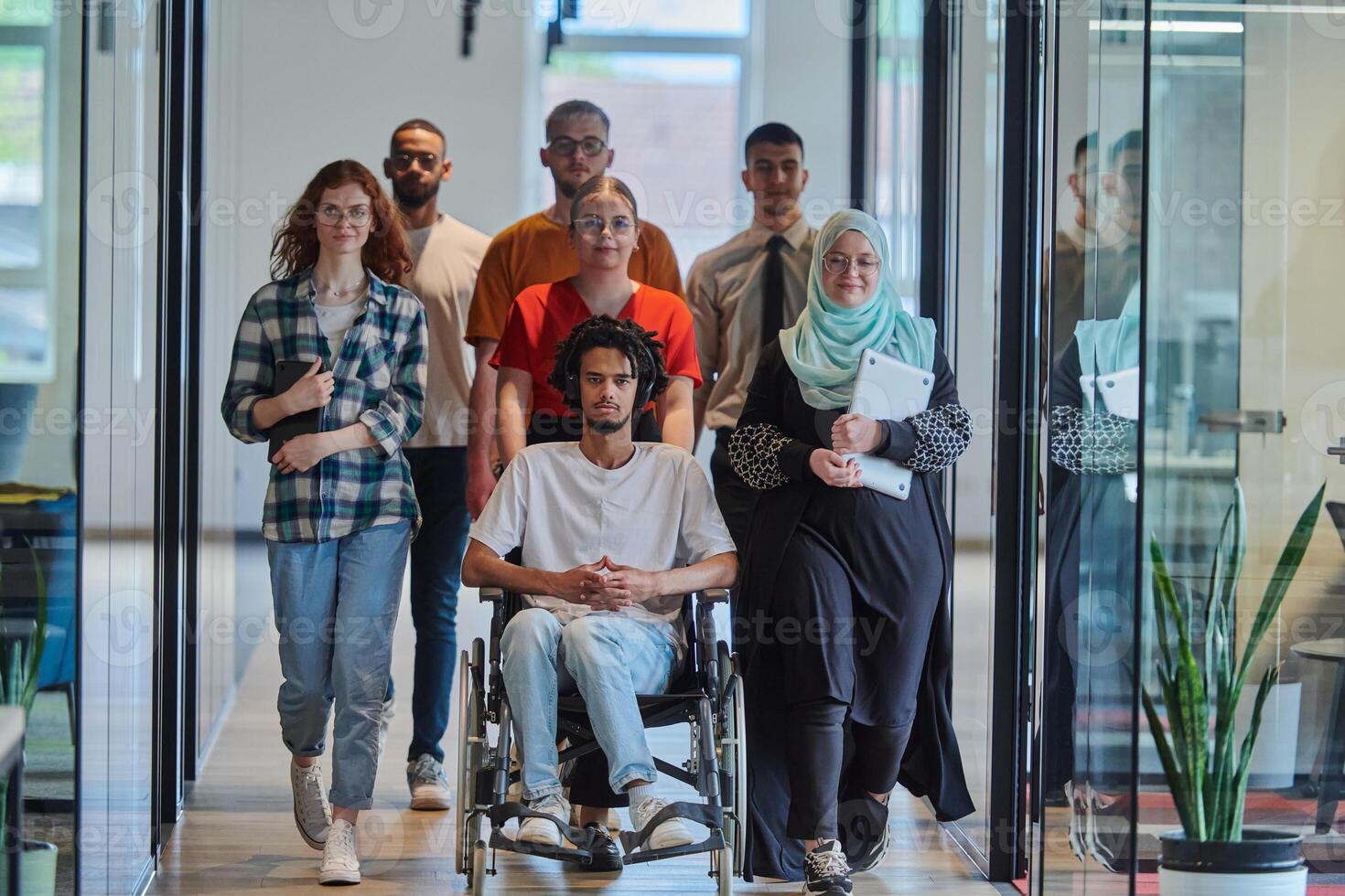 A diverse group of young business people walking a corridor in the glass-enclosed office of a modern startup, including a person in a wheelchair and a woman wearing a hijab, showing a dynamic mix of photo