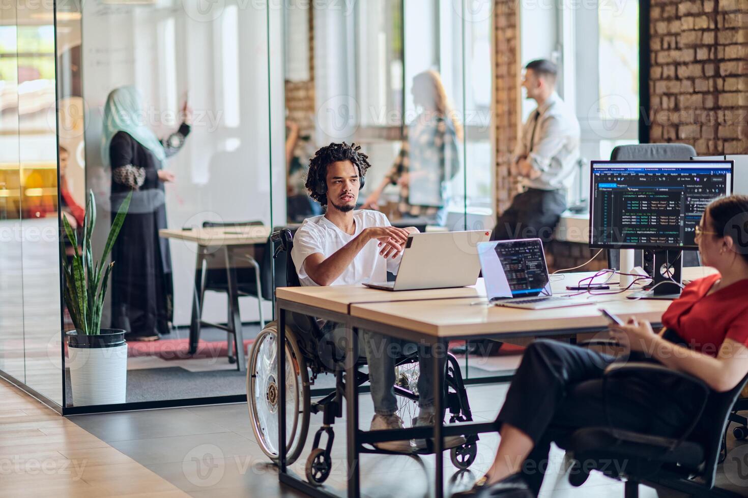 A young business group, including an African American businessman in a wheelchair, collaborates within a modern glass office, actively engaged around a computer and laptop, collectively solving photo