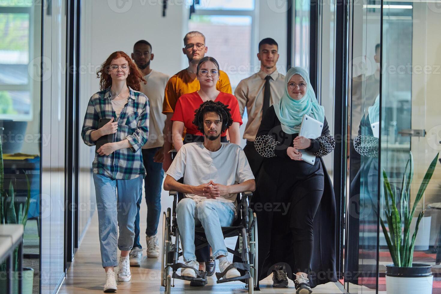 A diverse group of young business people walking a corridor in the glass-enclosed office of a modern startup, including a person in a wheelchair and a woman wearing a hijab, showing a dynamic mix of photo