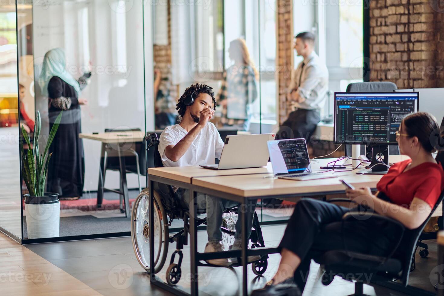 A young business group, including an African American businessman in a wheelchair, collaborates within a modern glass office, actively engaged around a computer and laptop, collectively solving photo