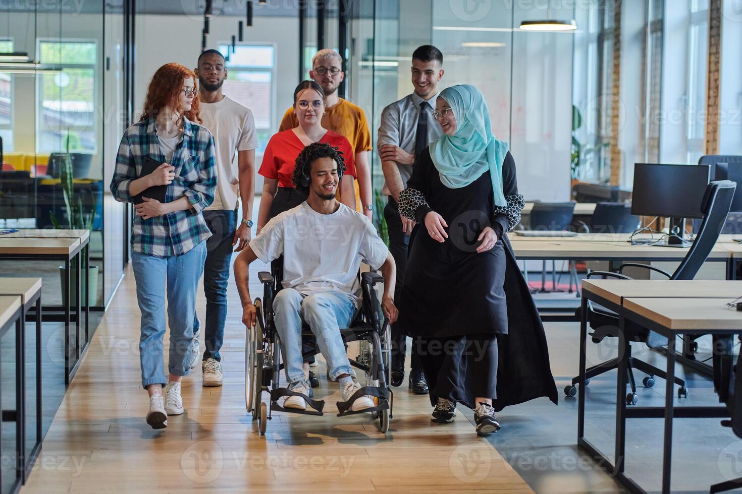 A diverse group of young business people walking a corridor in the glass-enclosed office of a modern startup, including a person in a wheelchair and a woman wearing a hijab, showing a dynamic mix of photo