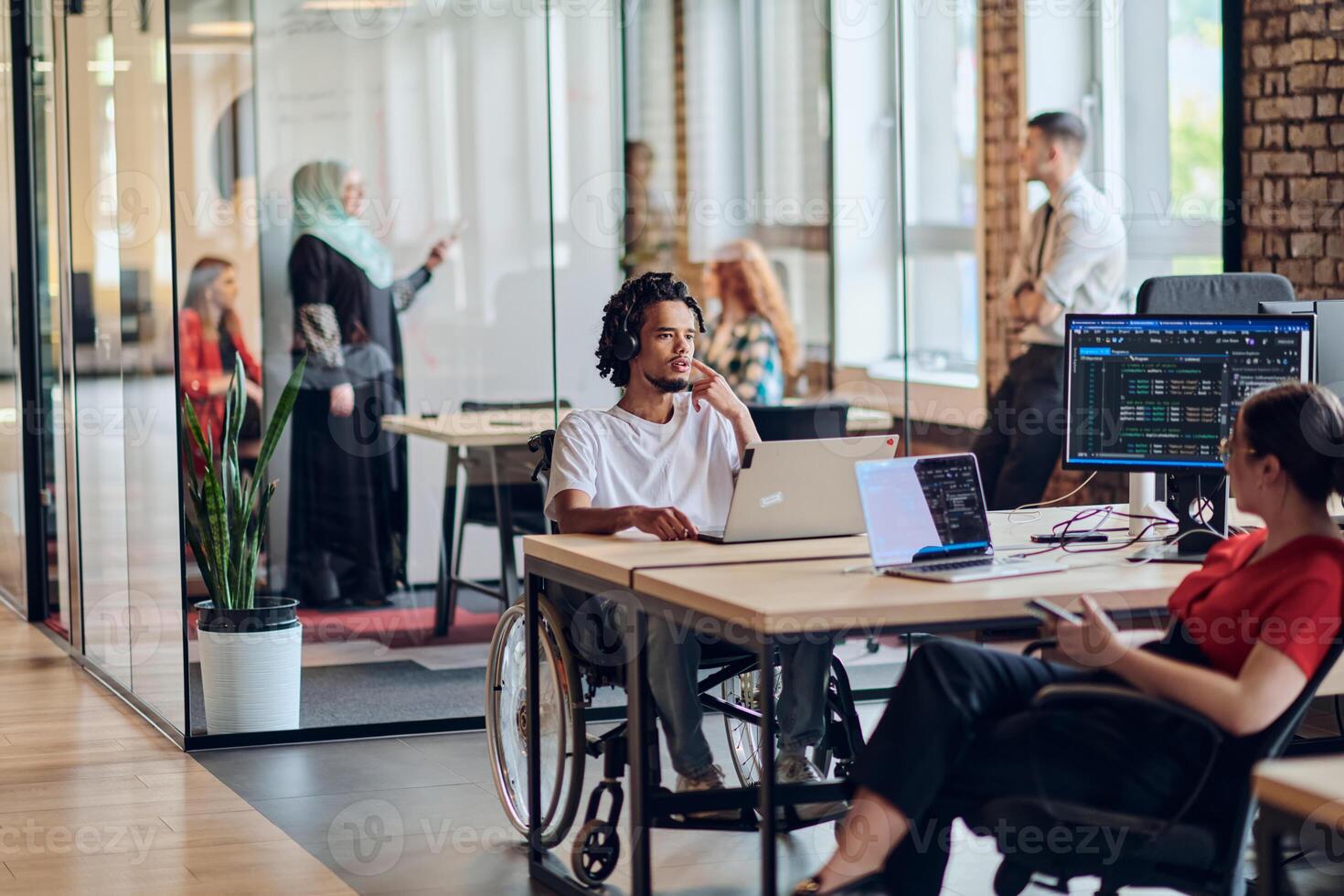 A young business group, including an African American businessman in a wheelchair, collaborates within a modern glass office, actively engaged around a computer and laptop, collectively solving photo