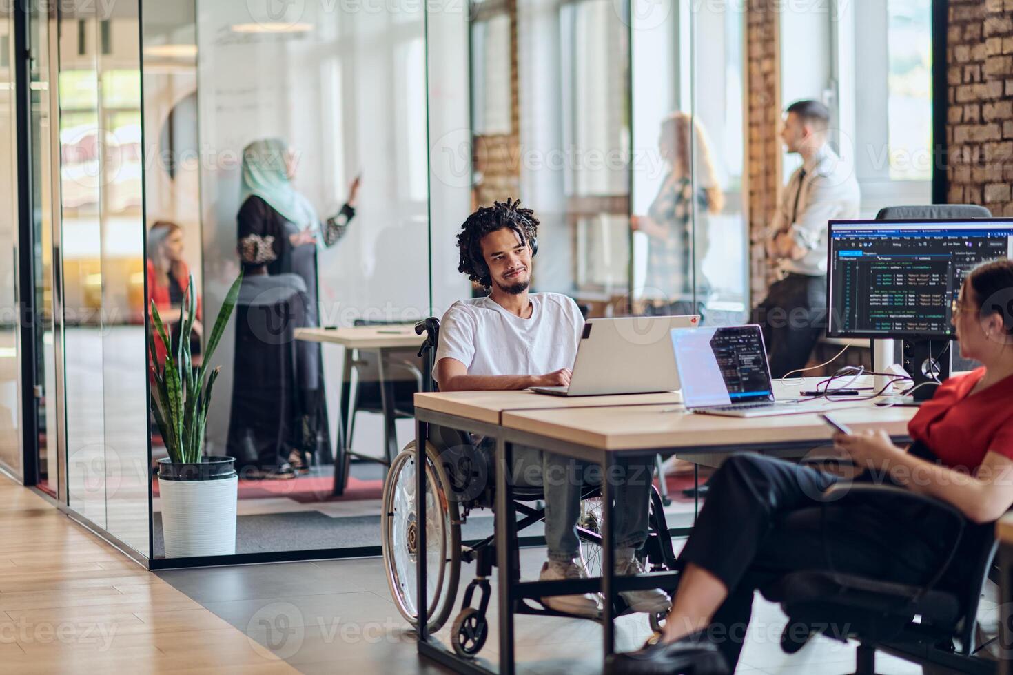 A young business group, including an African American businessman in a wheelchair, collaborates within a modern glass office, actively engaged around a computer and laptop, collectively solving photo