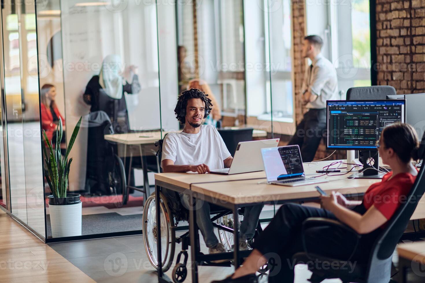 A young business group, including an African American businessman in a wheelchair, collaborates within a modern glass office, actively engaged around a computer and laptop, collectively solving photo