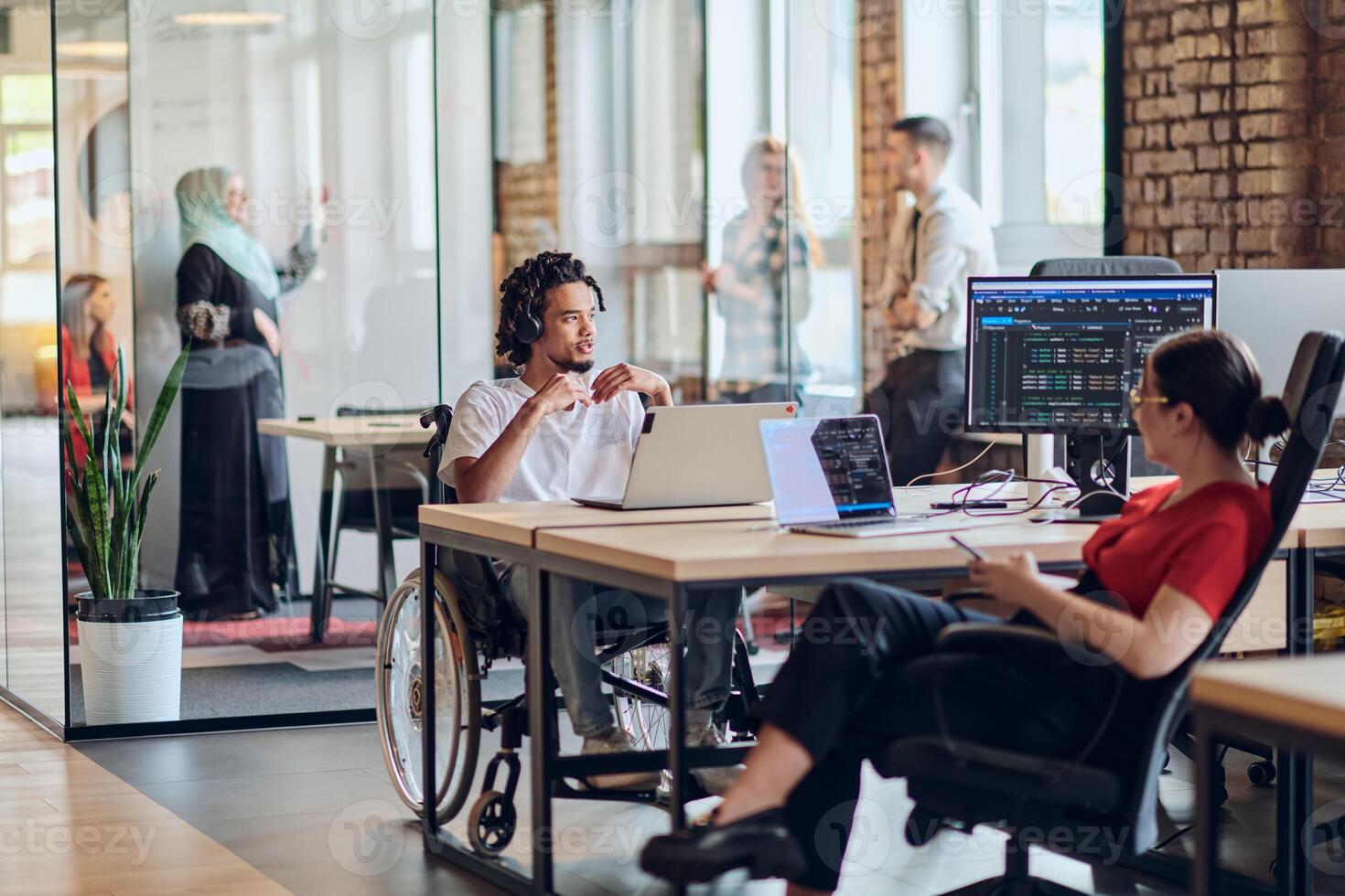A young business group, including an African American businessman in a wheelchair, collaborates within a modern glass office, actively engaged around a computer and laptop, collectively solving photo