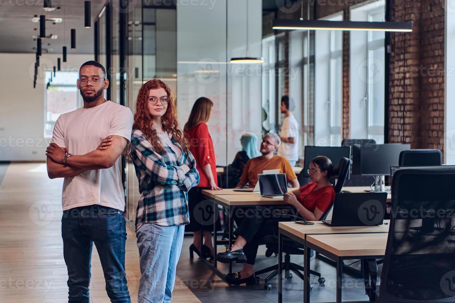A young African American businessman and a modern businesswoman with orange hair stand side by side, arms crossed, exuding confidence and unity in a contemporary office setting, epitomizing dynamic photo