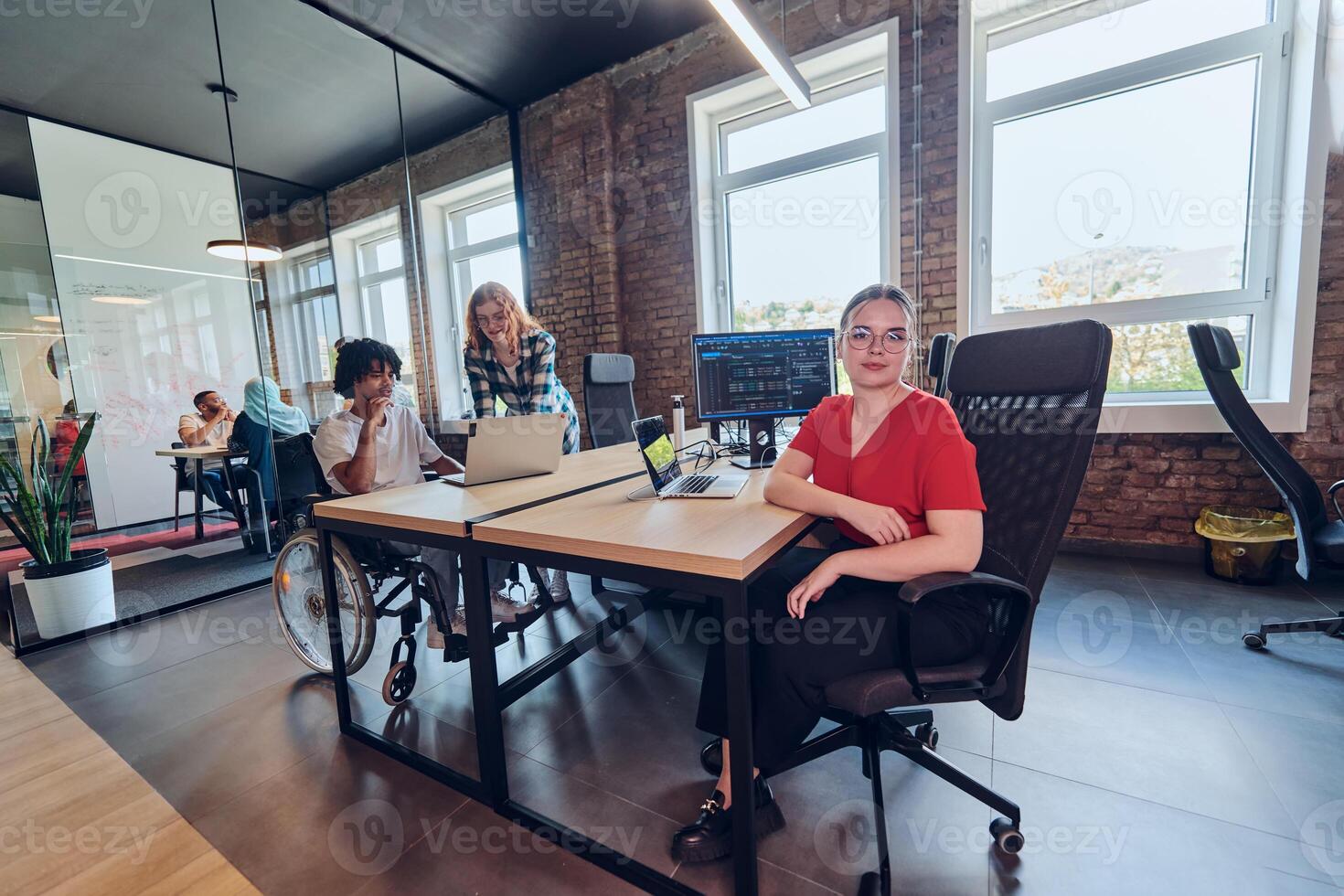 A young business group, including an African American businessman in a wheelchair, collaborates within a modern glass office, actively engaged around a computer and laptop, collectively solving photo