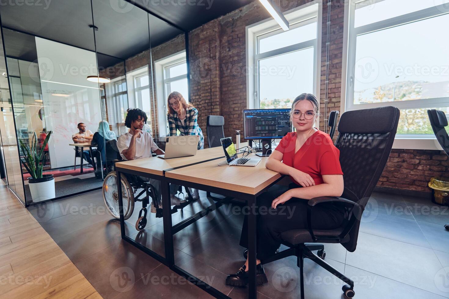 A young business group, including an African American businessman in a wheelchair, collaborates within a modern glass office, actively engaged around a computer and laptop, collectively solving photo