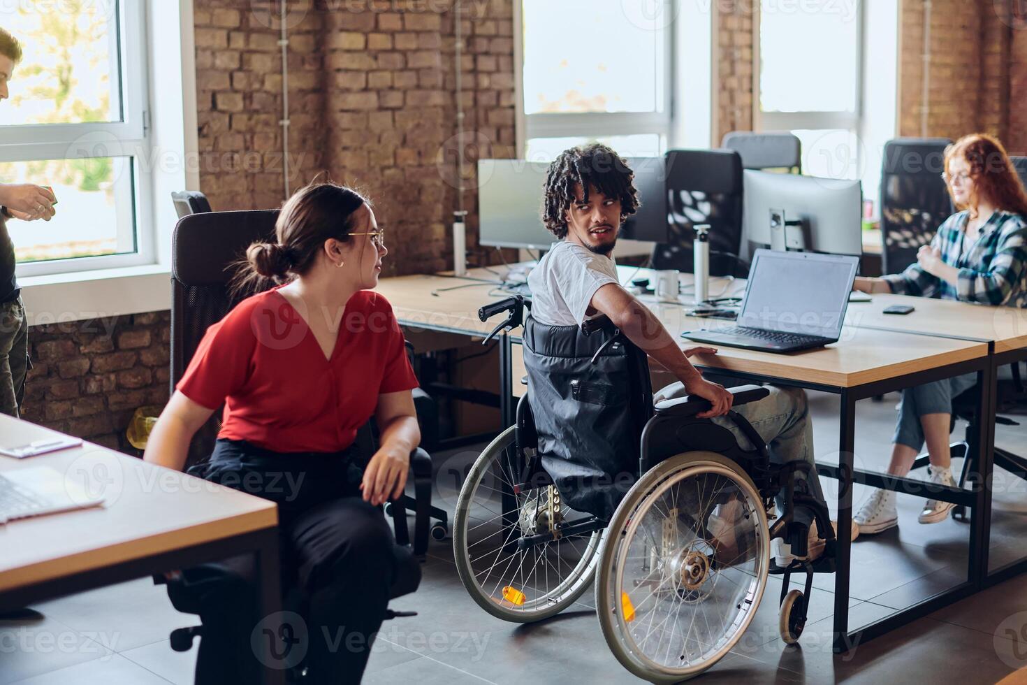 A young business group, including an African American businessman in a wheelchair, collaborates within a modern glass office, actively engaged around a computer and laptop, collectively solving photo