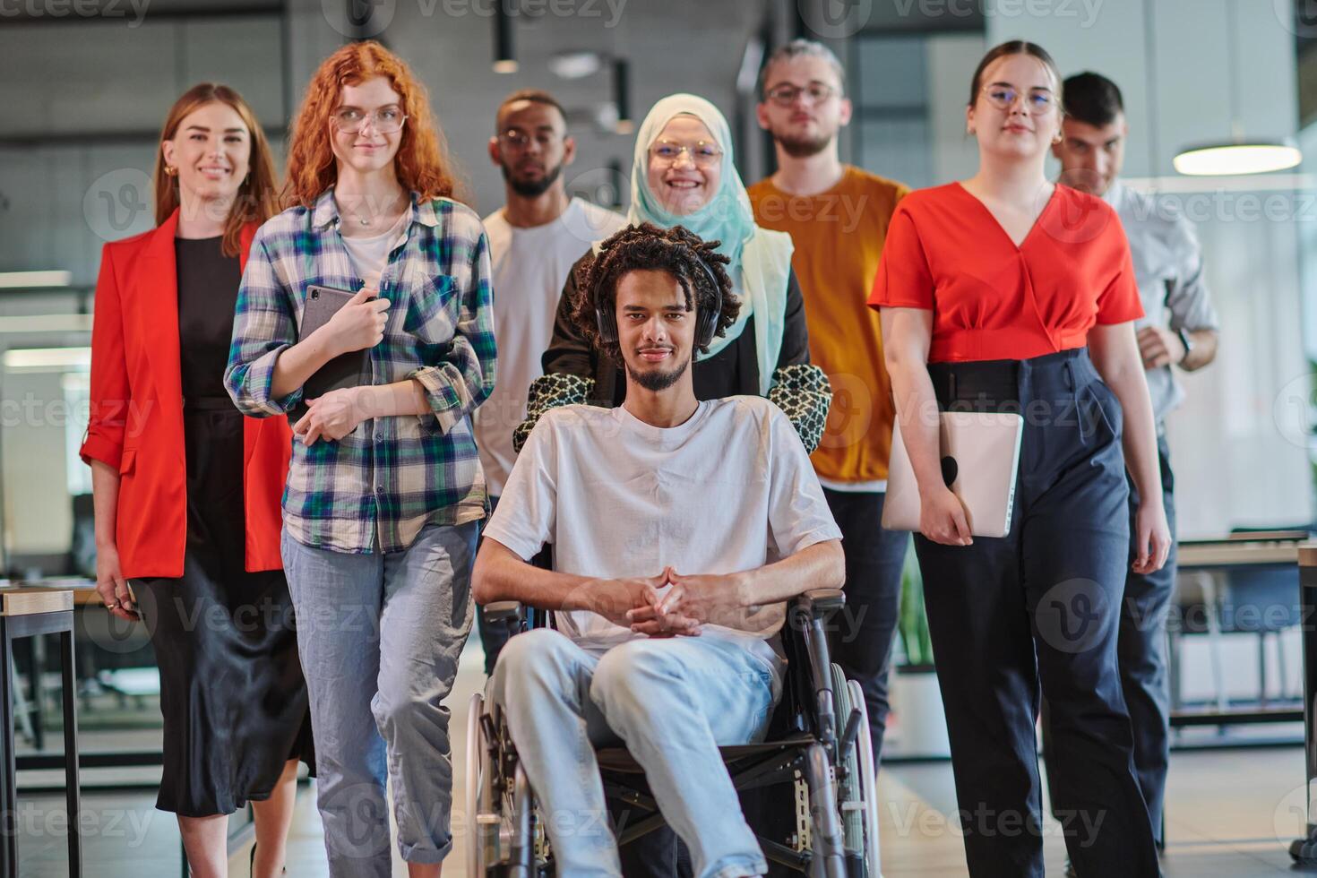 A diverse group of young business people congregates within a modern startup's glass-enclosed office, featuring inclusivity with a person in a wheelchair, an African American young man , and a hijab photo