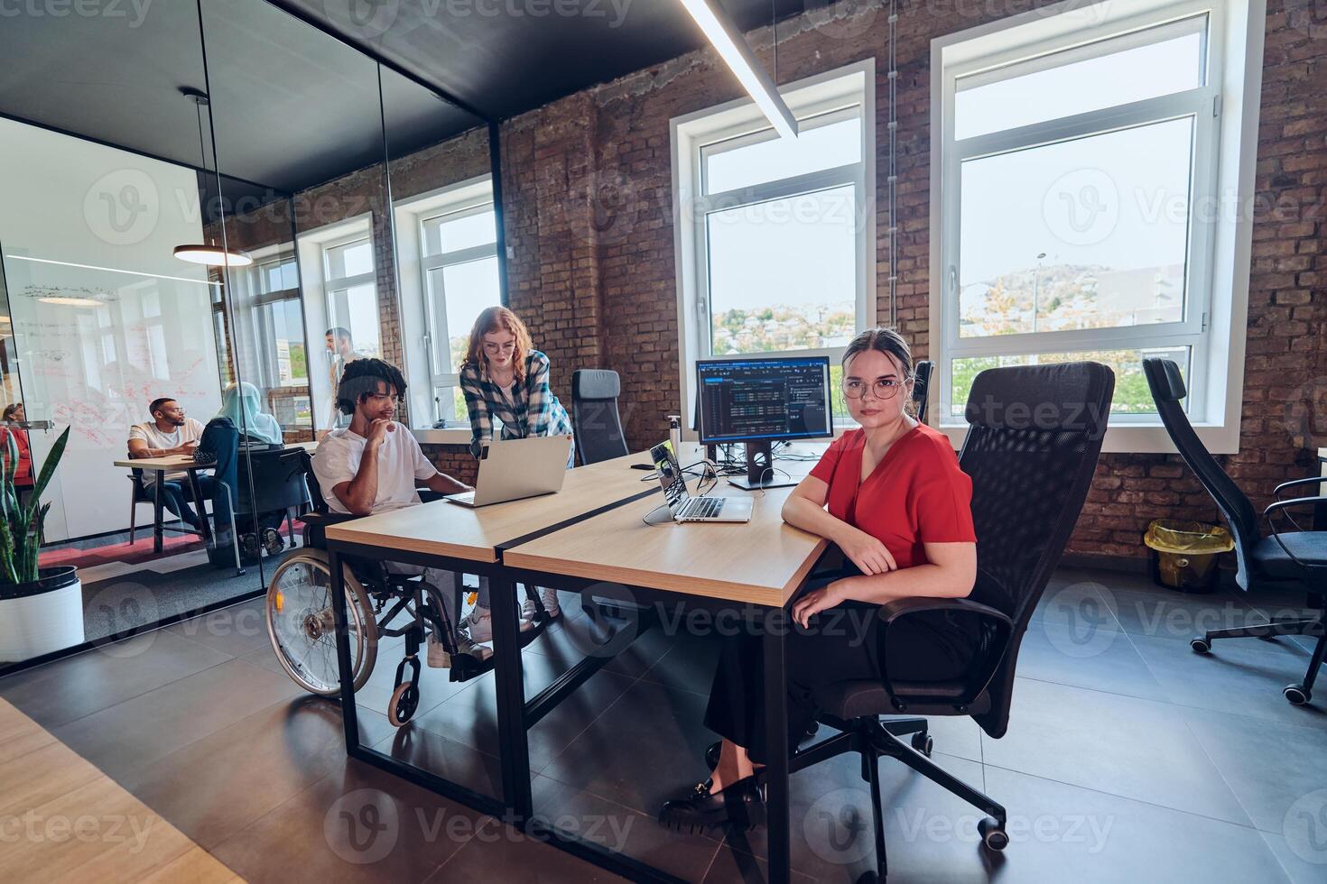 A young business group, including an African American businessman in a wheelchair, collaborates within a modern glass office, actively engaged around a computer and laptop, collectively solving photo