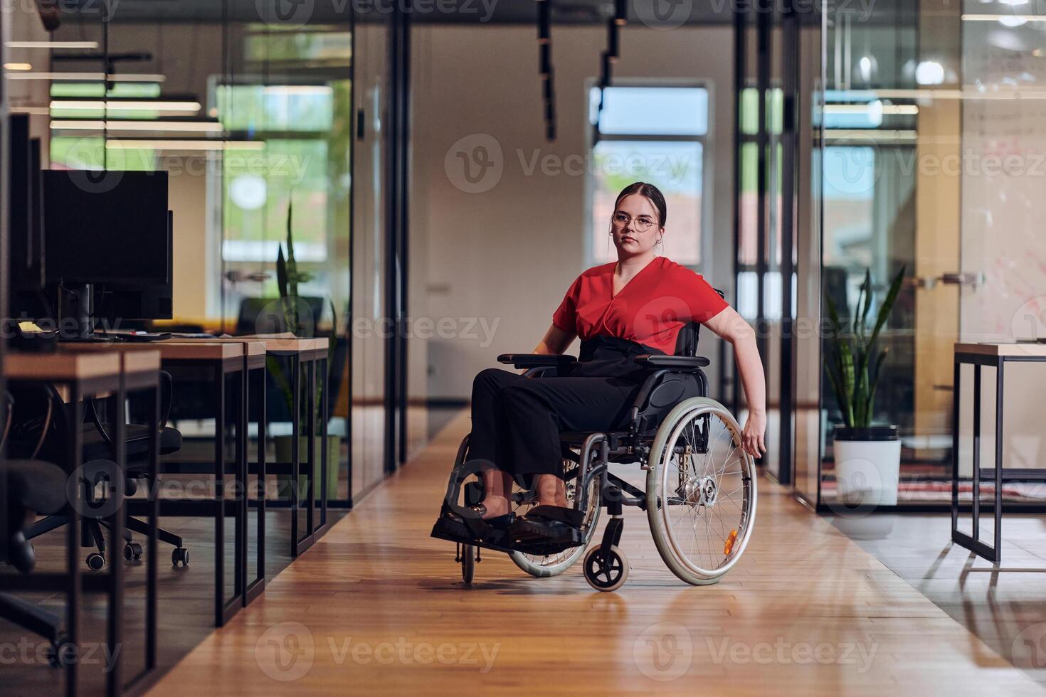 A modern young businesswoman in a wheelchair is surrounded by an inclusive workspace with glass-walled offices, embodying determination and innovation in the business world photo