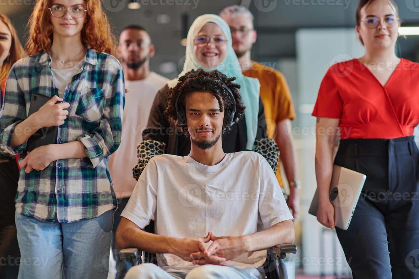 A diverse group of young business people congregates within a modern startup's glass-enclosed office, featuring inclusivity with a person in a wheelchair, an African American young man , and a hijab photo
