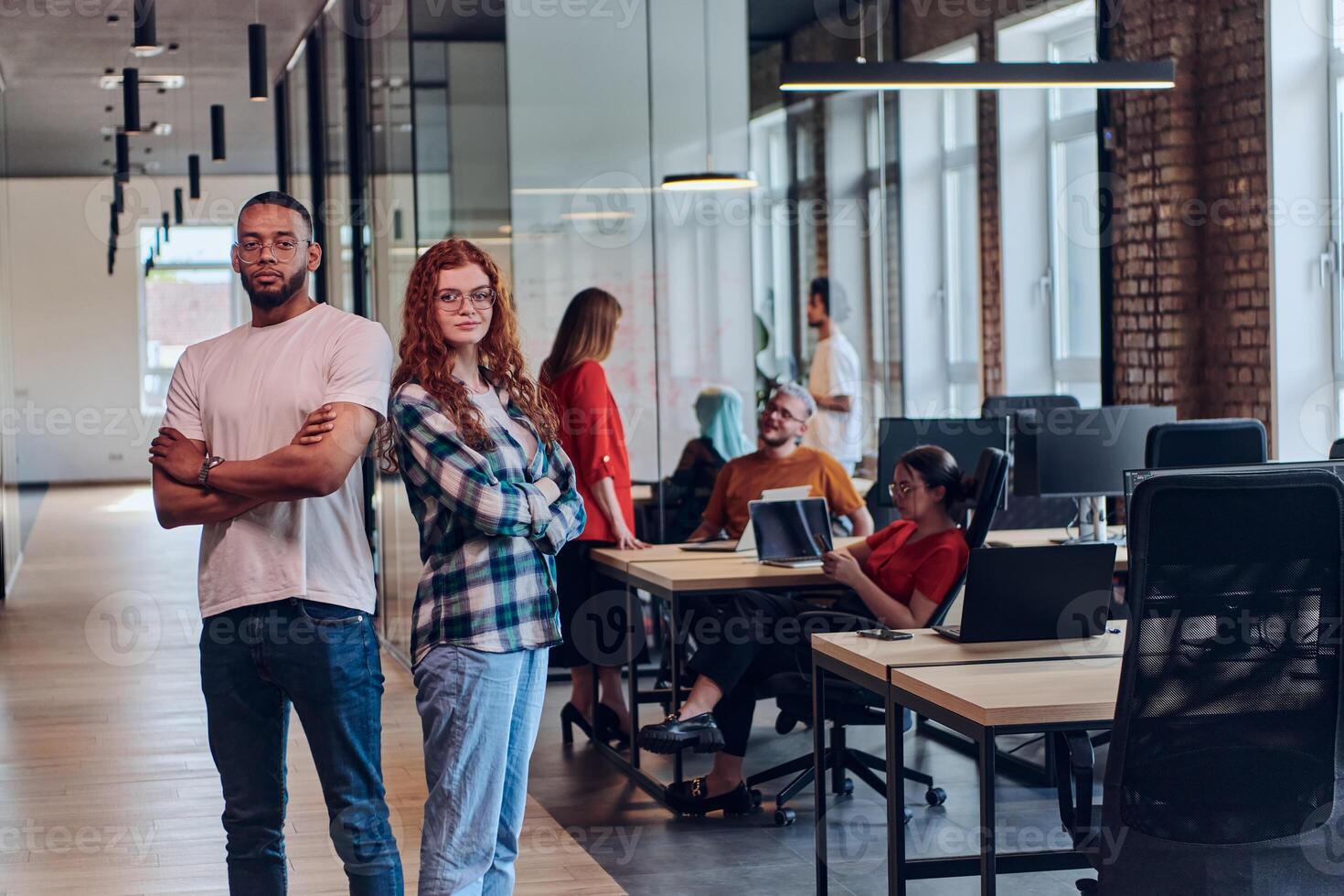 A young African American businessman and a modern businesswoman with orange hair stand side by side, arms crossed, exuding confidence and unity in a contemporary office setting, epitomizing dynamic photo