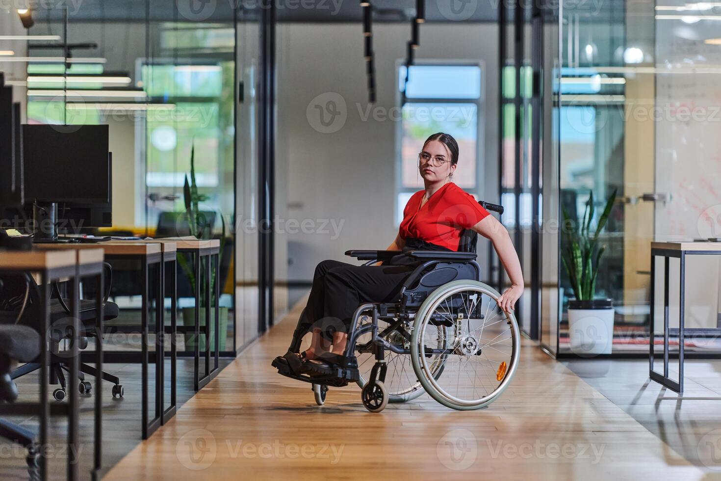 A modern young businesswoman in a wheelchair is surrounded by an inclusive workspace with glass-walled offices, embodying determination and innovation in the business world photo