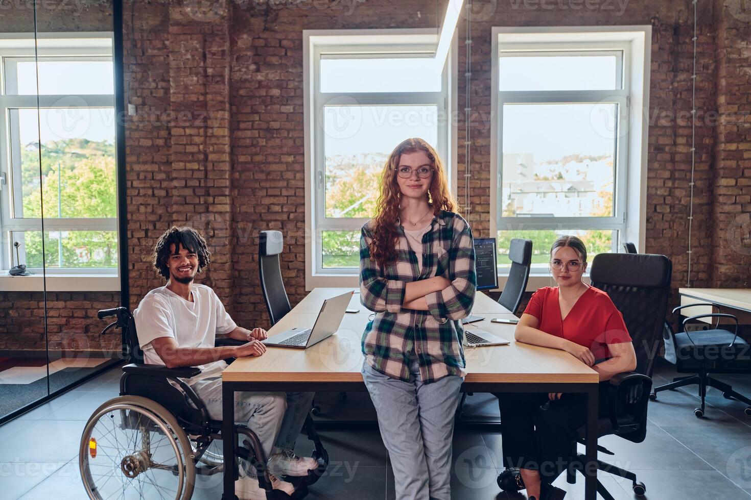 A young business group, including an African American businessman in a wheelchair, collaborates within a modern glass office, actively engaged around a computer and laptop, collectively solving photo