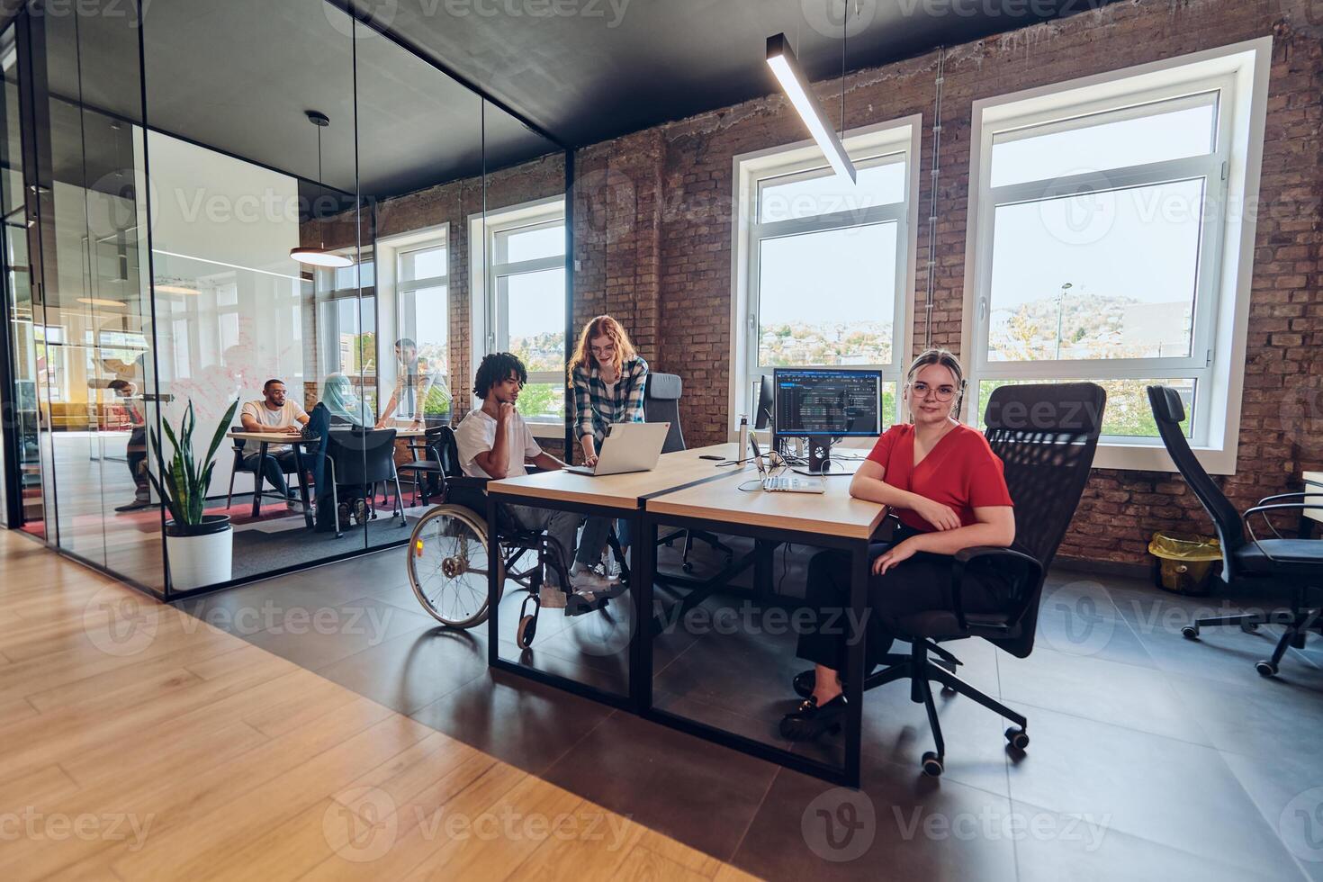 A young business group, including an African American businessman in a wheelchair, collaborates within a modern glass office, actively engaged around a computer and laptop, collectively solving photo