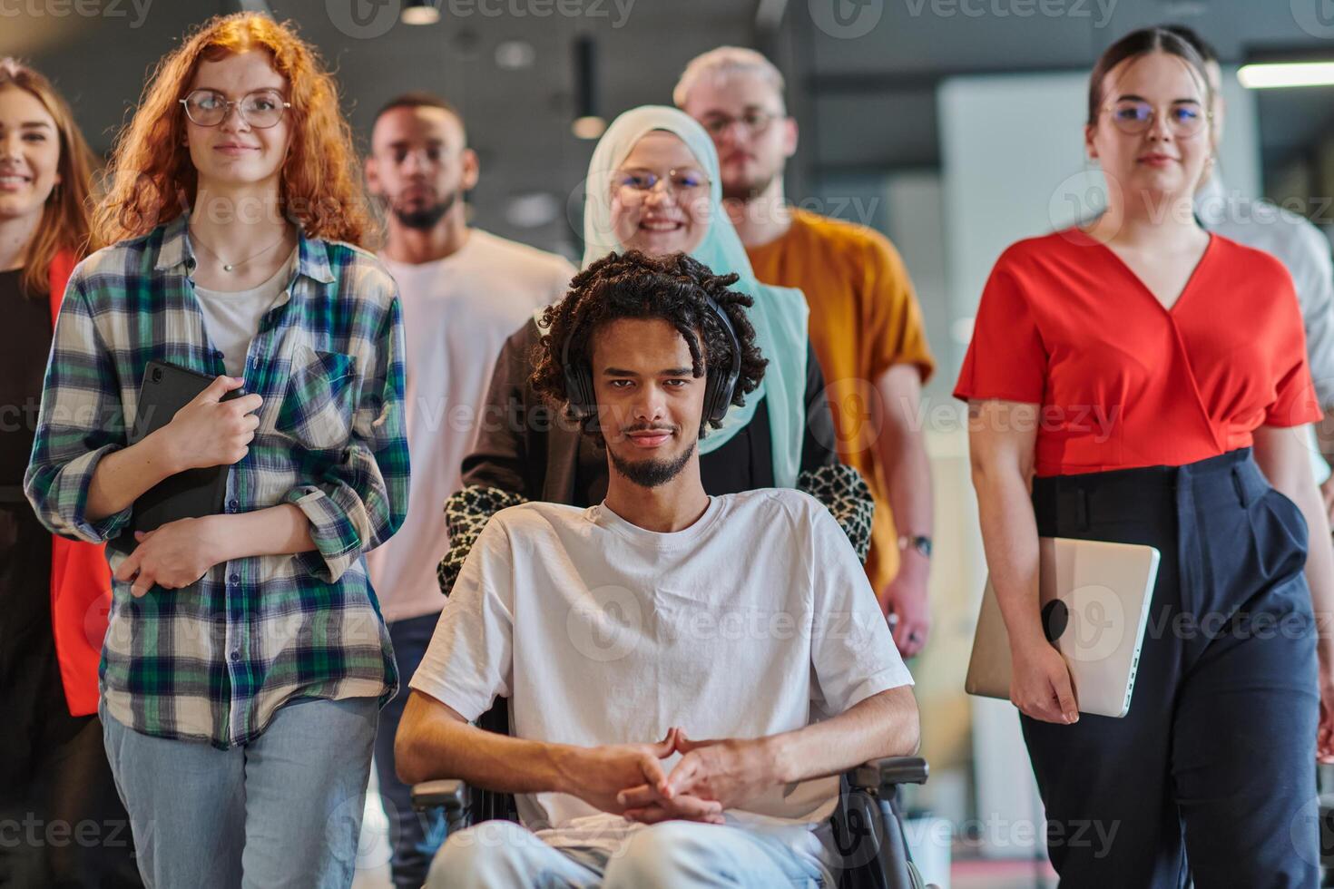 A diverse group of young business people congregates within a modern startup's glass-enclosed office, featuring inclusivity with a person in a wheelchair, an African American young man , and a hijab photo
